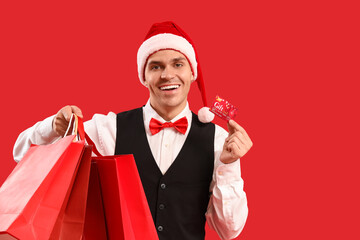 Poster - Handsome young happy man in Santa hat with gift card and shopping bags on red background