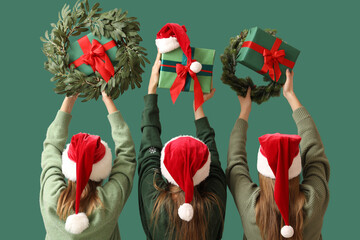 Wall Mural - Women in Santa hats holding Christmas gift boxes and wreathes on green background, back view