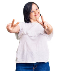 Sticker - Brunette woman with down syndrome wearing casual white tshirt smiling with tongue out showing fingers of both hands doing victory sign. number two.