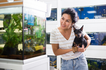 Wall Mural - Positive asian woman with dog in her arms choosing aquarium fish at pet shop