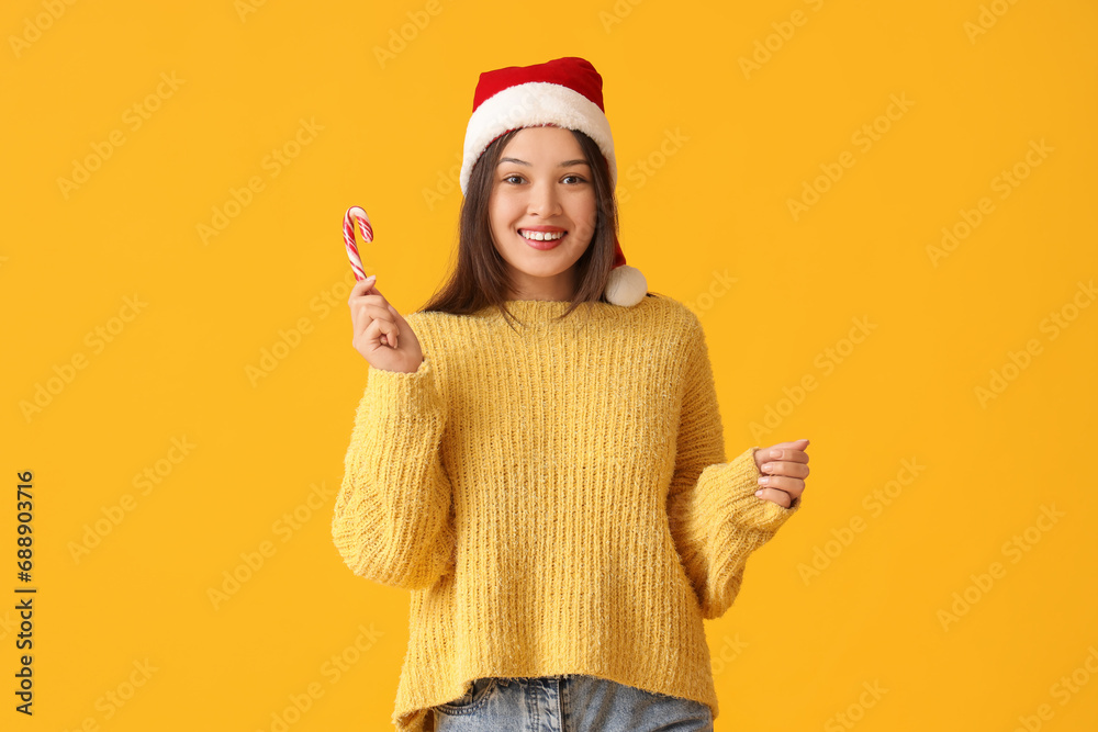 Happy young Asian woman in Santa hat with candy cane on yellow background