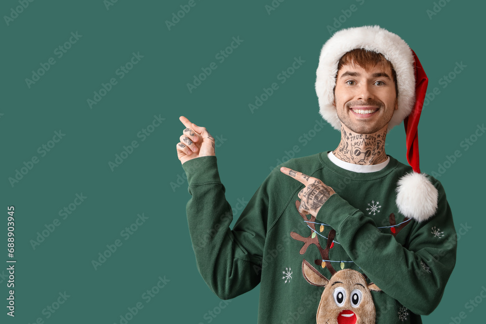 Tattooed young man in Santa hat pointing at something on green background