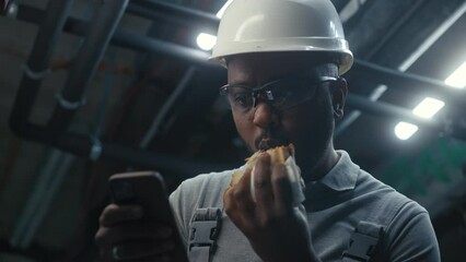 Wall Mural - African American engineer eats sandwich and surfs the Internet on phone. Professional worker in uniform and protective hard hat having break working on industrial factory or energy facility. Close up.