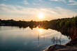 This image captures the serene atmosphere of a quarry lake at sunset. The sun hovers just above the treeline, casting a soft glow over the still water. The lake, flanked by rocky edges and sparse
