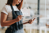 Fototapeta  - Happy excited attractive young Asian woman in denim apron, received online order in coffee shop with an 'Open' sign.