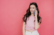 Satisfied brunette girl talking by phone toothy smiling standing against pink studio background looks at  copy space. Promo, sale, gossip concept.