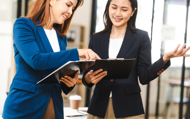 Two middle aged and young Asian female executives in formal suits review bar chart, discussing business strategies in office setting, senior executives or directors in advertising or public relations