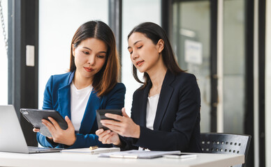 Two middle aged and young Asian female executives in formal suits review bar chart, discussing business strategies in office setting, senior executives or directors in advertising or public relations