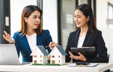 Two Asian real estate agents in formal suits discuss a contract with house models on the table, ready to sign a deal.