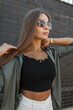 © alones - Fashionable beautiful young woman model with sunglasses with a black top and stylish shirt stands near a black brick wall. Pretty chic lady with urban outfit