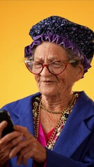 Wall Mural - Vertical video of toothless funny old grandmother wearing glasses and boa having video chat with family and friends isolated on yellow background. Concept of crazy senior youthful woman.