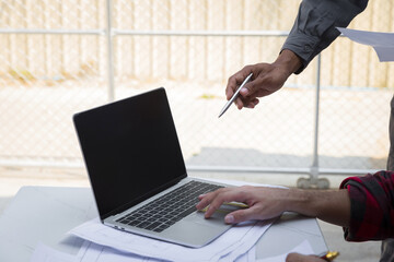 Wall Mural - Engineers and architects pointing at laptop screens looking at blueprints. Two Construction Engineers or Architects discussing at work with laptop on construction site and pointing on laptop.