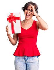 Sticker - Beautiful young woman with short hair holding gift smiling happy doing ok sign with hand on eye looking through fingers