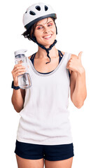 Poster - Beautiful young woman with short hair wearing bike helmet and holding water bottle smiling happy and positive, thumb up doing excellent and approval sign