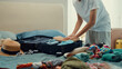 © tirachard - Young happy and exciting Asian female ready for summer holidays prepare suitcase put cloth on her bag in full of messy stuff bedroom. Preparing luggage for summer vacation trip. Travel concept.