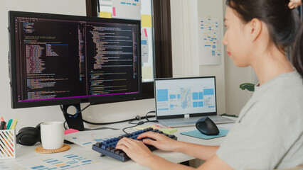 Close-up young Asia girl IT development programmer typing on keyboard coding programming fixing database code on computer monitor and laptop screen on table in workroom at house office.