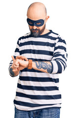 Wall Mural - Young handsome man wearing burglar mask checking the time on wrist watch, relaxed and confident