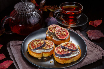 Wall Mural - Toast with cream cheese, fig slices, pine nuts, sunflower seeds and sesame seeds on a plate. Hot tea in a cup and teapot on a dark table