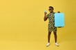 Happy handsome black man in glasses holding blue travel suitcase high in hand reading message on phone. On yellow background stands positive stylish African with mobile phone and suitcase in hand