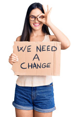 Canvas Print - Young beautiful caucasian woman holding we need a change banner smiling happy doing ok sign with hand on eye looking through fingers