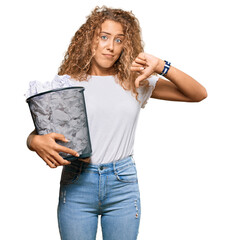 Sticker - Beautiful caucasian teenager girl holding paper bin full of crumpled papers with angry face, negative sign showing dislike with thumbs down, rejection concept