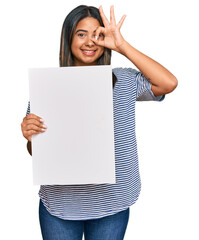 Poster - Young latin girl holding blank empty banner smiling happy doing ok sign with hand on eye looking through fingers