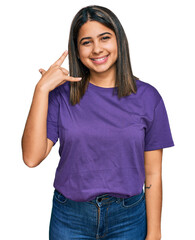 Poster - Young hispanic girl wearing casual purple t shirt smiling doing phone gesture with hand and fingers like talking on the telephone. communicating concepts.