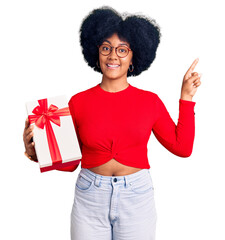 Sticker - Young african american girl holding gift with a big smile on face, pointing with hand finger to the side looking at the camera.