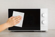 Young adult woman hand holding dry white paper napkin and wiping dark black glass door surface of white microwave oven on table top at home kitchen. Closeup. Regular cleanup. Front view.