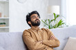 © Liubomir - Tired young Indian man napping on couch at home with arms folded on chest, hard day's work, rest.
