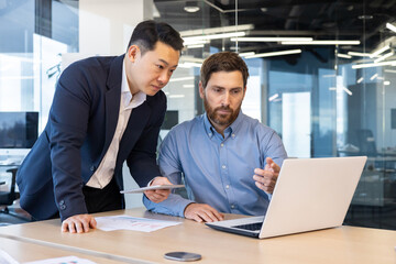 Two businessmen in suits work in the office, look at a laptop, discuss work issues, have a discussion.
