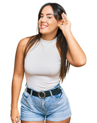 Young beautiful hispanic girl wearing casual clothes smiling with hand over ear listening an hearing to rumor or gossip. deafness concept.