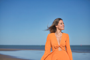 Wall Mural - Young and beautiful woman in an orange dress walking on the beach, happy and relaxed, with the wind moving her dress and hair. Concept beauty, fashion, trend.