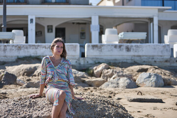 Wall Mural - Young and beautiful woman in a sequined dress and high heels, sitting on a large rock on the beach, happy, relaxed and calm. Concept beauty, fashion, trend, peace, relaxation.