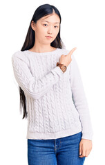 Wall Mural - Young beautiful chinese woman wearing casual sweater pointing with hand finger to the side showing advertisement, serious and calm face