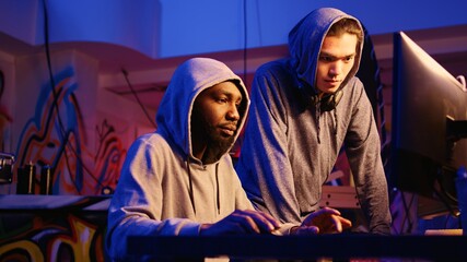 Poster - African american hacker talking to asian colleague in rundown hiding place, brainstorming ways to find exploits for system vulnerabilities and use them to ask system owner for ransom