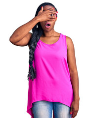 Poster - Young african american woman wearing casual clothes and glasses peeking in shock covering face and eyes with hand, looking through fingers with embarrassed expression.