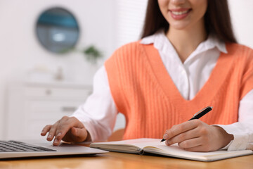 Canvas Print - Young woman writing in notebook while working on laptop at wooden table, closeup