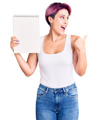 Canvas Print - Young beautiful woman with pink hair holding notebook pointing thumb up to the side smiling happy with open mouth