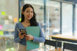 © Songsak C - Young beautiful Asian business woman holding a folder and a smart phone while standing at the workplace.