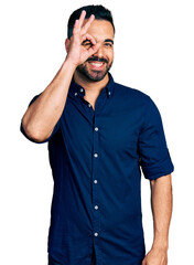 Poster - Young hispanic man with beard wearing casual blue shirt smiling happy doing ok sign with hand on eye looking through fingers