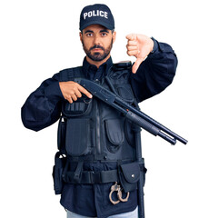 Wall Mural - Young hispanic man wearing police uniform holding shotgun with angry face, negative sign showing dislike with thumbs down, rejection concept