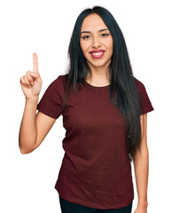 Wall Mural - Young hispanic girl wearing casual t shirt showing and pointing up with finger number one while smiling confident and happy.