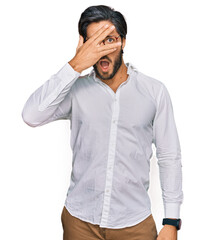 Wall Mural - Young hispanic man wearing business shirt and glasses peeking in shock covering face and eyes with hand, looking through fingers with embarrassed expression.