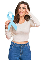 Canvas Print - Young brunette woman holding blue ribbon stressed and frustrated with hand on head, surprised and angry face