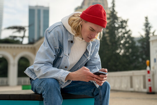 Stylish hipster man sitting in urban street with smartphone, reading, messaging, searching information, posting photos in social networks. Slim guy looks like blogger, tourist, international student
