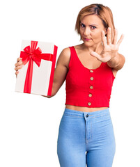 Sticker - Young blonde woman holding gift with open hand doing stop sign with serious and confident expression, defense gesture