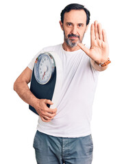 Poster - Middle age handsome man holding weighing machine with open hand doing stop sign with serious and confident expression, defense gesture