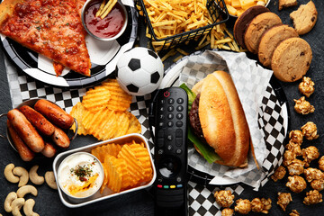 Wall Mural - Saturated fats. Football time. TV remote control and snacks - chips, popcorn, cookies, cheese, sauce, fries, burger, nuts