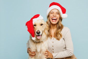 Wall Mural - Young joyful owner woman wears casual clothes Santa hat hug cuddle embrace best friend pet retriever dog wink isolated on plain pastel blue background studio. New Year Christmas celebration concept.